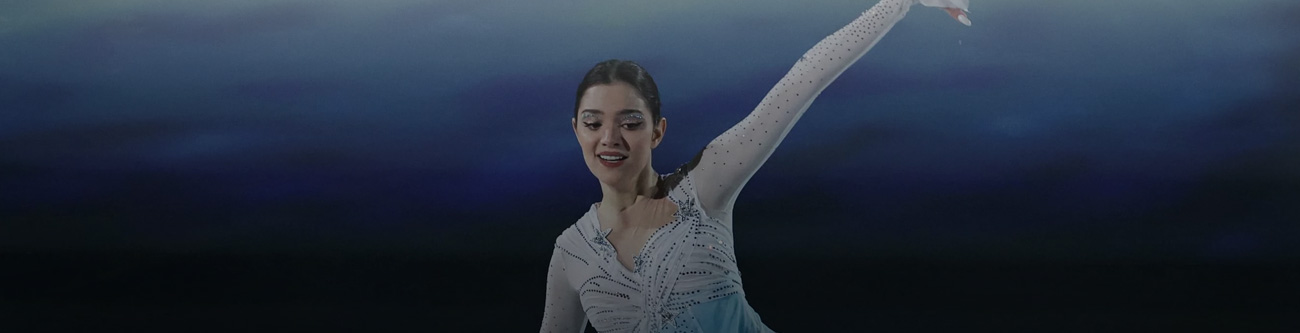 Изображение девушки в костюме фигуриста, выполняющей элемент на льду. Она одета в костюм с длинными рукавами, украшенный блестящими деталями. Фон затемнён и напоминает разноцветные тени или ледовую арену